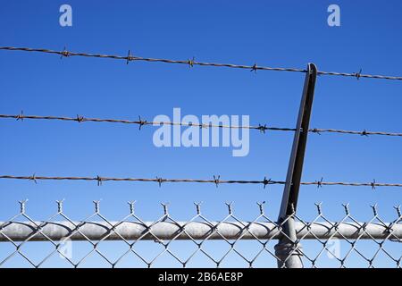 barbed wire angled over the top of a chain link fence with blue sky background Stock Photo