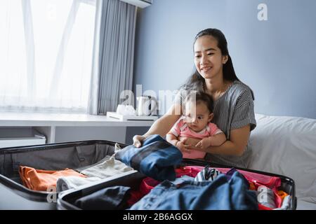 asian mother sits on her baby girl's lap while preparing clothes to put in the suitcase