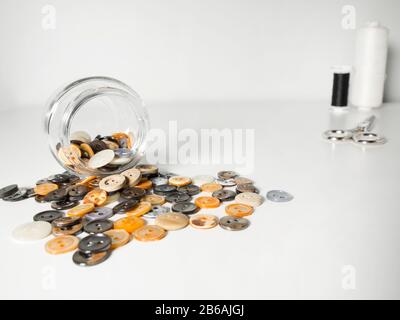 Glass jar full of clothing orange, yellow and gray buttons, thread and scissors on white background. Sewing and craft concept. Stock Photo