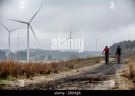 Two men ride mountain bikes in the shadow of wind turbines near Lluest-Wen Reservoir in South Wales. Stock Photo