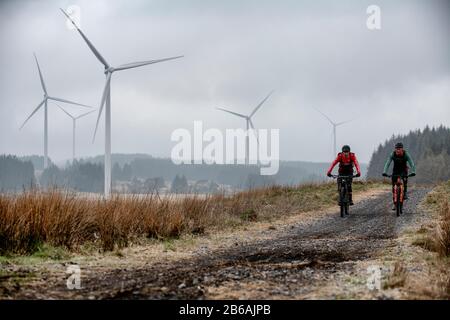 Two men ride mountain bikes in the shadow of wind turbines near Lluest-Wen Reservoir in South Wales. Stock Photo