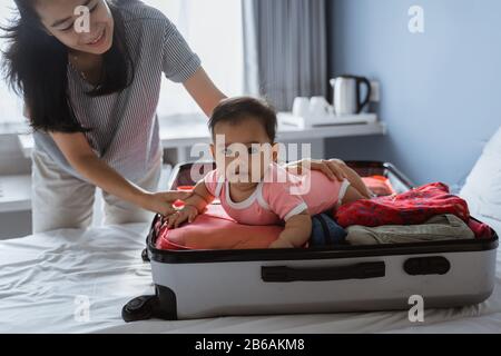 Mom smiles holding a cute baby lying in an open suitcase filled with clothes placed on the bed