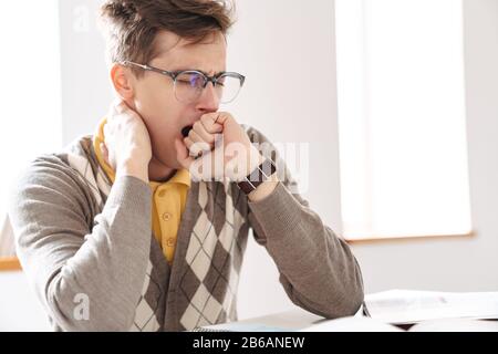 Picture of a young tired guy student sit at the table indoors yawning. Stock Photo