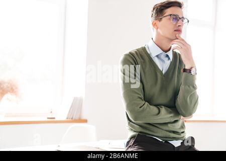 Image of a thinking thoughtful young guy student sit on table indoor in eyeglasses. Stock Photo