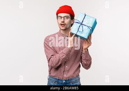 Waist-up portrait of curious funny caucasian bearded man in red beanie, glasses, shaking gift box near ear as trying guess whats inside, look focused Stock Photo