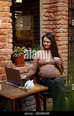 Portrait of young pregnanty woman freelancer using laptop computer for distance job while sitting in modern coffee with cup of tea Stock Photo