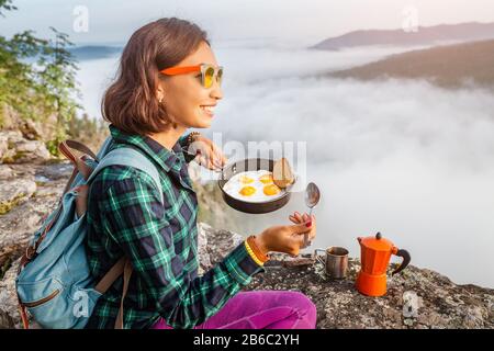 A woman traveler cooking and eating breakfast scrambled eggs and coffee at dawn in the misty mountains. Camping equipment and adventure concept Stock Photo