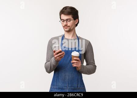 Portrait of carefree handsome young male student working part-time in restaurant in apron, have lunch break, using mobile phone and drink coffee form Stock Photo