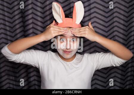 A cute and happy girl smears face with cosmetic clay or mud, standing in the bathroom, with rabbit ears on her head and smiling. The concept of health and beauty. Stock Photo