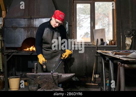 woman blacksmith metal artist forges a figured metal blank with hammer on the anvil in a traditional workshop Stock Photo