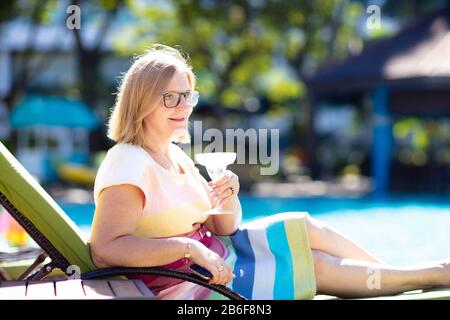 Woman drinking cocktail at swimming pool. Senior lady with exotic drink in luxury beach resort. Early retirement. Active retiree travel the world. Stock Photo