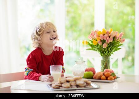 Child eating breakfast. Kid drinking milk with fresh homemade cookies. Baking and cooking with kids. Little boy with fruit bowl and pastry for family Stock Photo