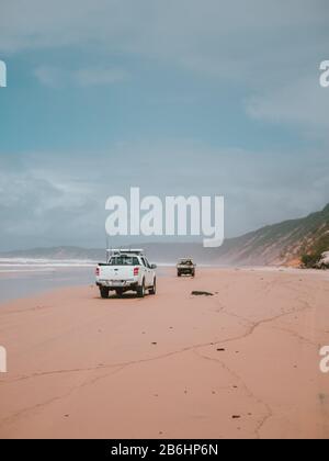 Two cars driving on the shore of the beach with mountains in the background in Australia Stock Photo
