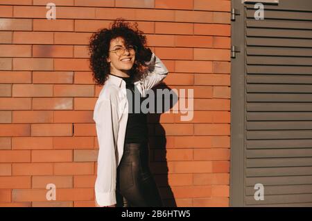 Caucasian businesswoman with nice curly hair and eyeglasses is posing cheerfully on a stone wall Stock Photo