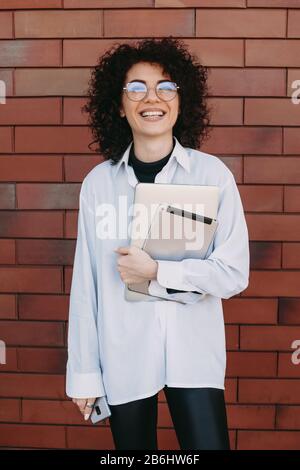 Lovely caucasian lady with curly hair and modern gadgets is posing on a stone wall while smiling