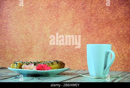 A high white mug with tea and a plate with three fresh eclairs in icing stand on a wooden table. Close-up. Stock Photo