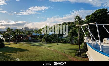 The bow of a boat leading the view out to a tropical Australian resort on the coast with a cloudy sky in early morning light Stock Photo