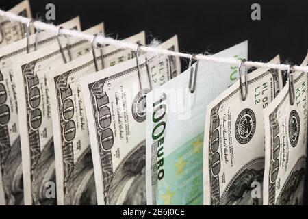 Parts of hundred dollar bills and one hundred euro bill between them is hanging on clerical clips on a rope on the dark background. Closeup, selective Stock Photo