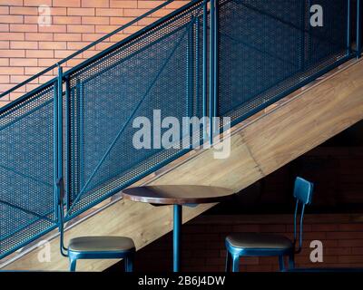 Cafe decoration design. Empty round wooden table with two modern bar stools near metal stairs on brick wall background. Stock Photo