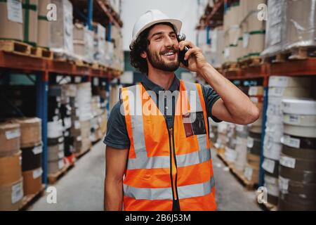 Smiling warehouse manager in conversation on mobile phone wearing white helmet and safety vest standing in between goods shelf looking away Stock Photo