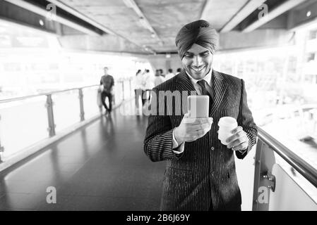 Happy young Indian Sikh businessman using phone at sky train station Stock Photo