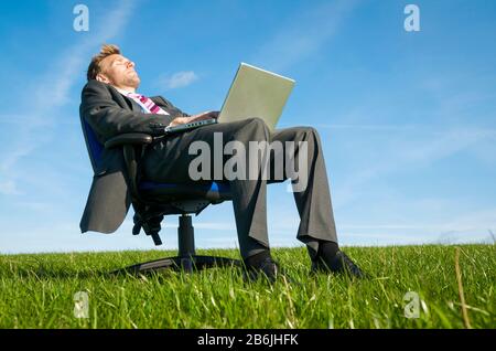 Relaxed businessman sitting outdoors on an office chair falling asleep at his laptop computer in a sunny green meadow Stock Photo