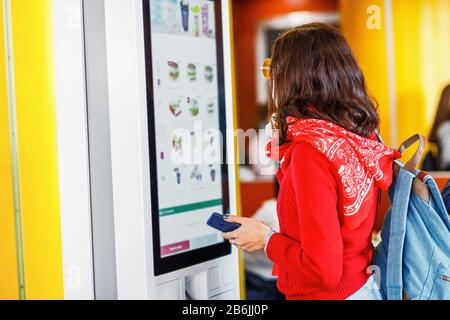 Hipster asian woman orders food at the touch screen self service machine by the electronic menu in the fastfood restaurant Stock Photo