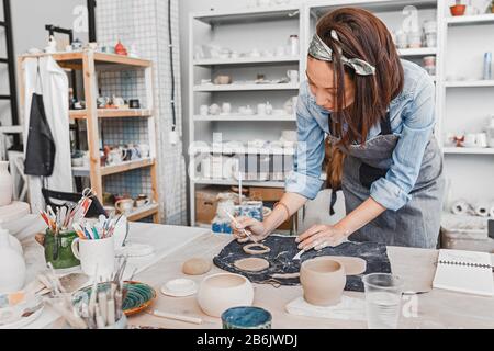 Close-up view of a female potter making and molding clay dishware in a workshop using different tools Stock Photo