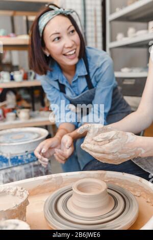 Two girls friends smiling and talking while working on potters wheel making clay handmade craft in pottery workshop, friendship and guidance concept Stock Photo