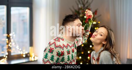 people and holiday traditions concept - portrait of happy couple in ugly sweaters kissing under mistletoe over home and christmas tree lights on backg Stock Photo