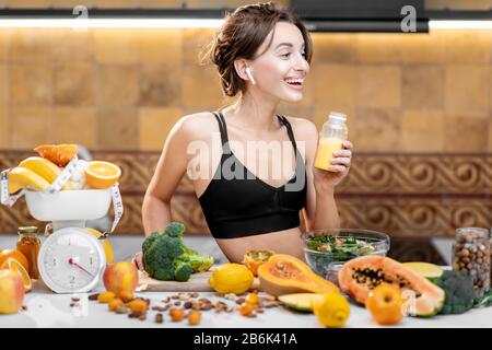Portrait of an athletic woman having a break, drinking juice while standing with lots of healthy fresh food on the kitchen. Concept of losing weight, sports and healthy eating