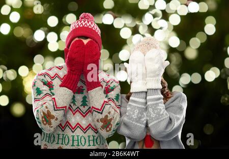 christmas, people and holidays concept - couple in ugly sweaters, hats and woolen mittens covering their faces over festive lights on dark green backg Stock Photo