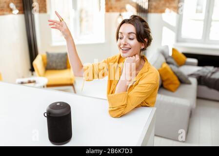 Happy young woman controlling smart home devices with a voice commands and smart speaker at home. Concept of a smart home and managing wireless devices remotely Stock Photo