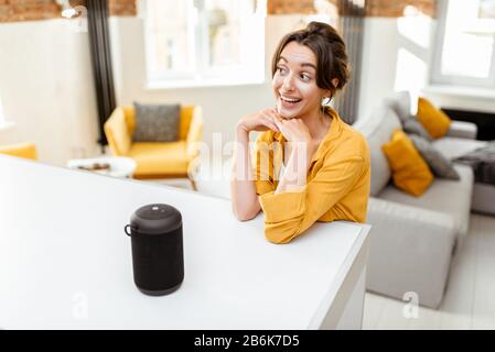 Happy young woman controlling smart home devices with a voice commands and smart speaker at home. Concept of a smart home and managing wireless devices remotely Stock Photo