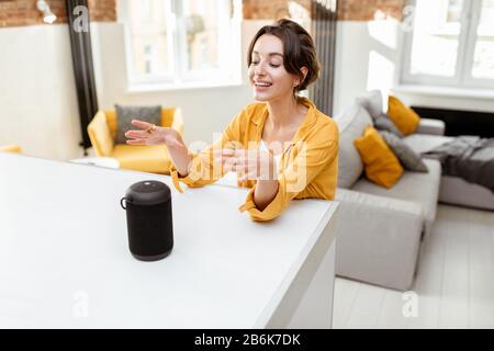 Happy young woman controlling smart home devices with a voice commands and smart speaker at home. Concept of a smart home and managing wireless devices remotely Stock Photo