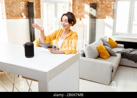 Happy young woman controlling smart home devices with a voice commands and smart speaker at home. Concept of a smart home and managing wireless devices remotely Stock Photo