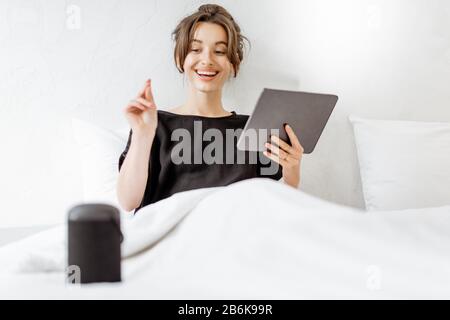 Young woman lying in bed with a digital tablet and smart speaker, controlling home devices with a voice commands. Concept of a smart home and relaxation at home Stock Photo