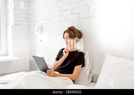 Young woman using digital tablet while sitting relaxed in the bed of a white bedrrom. Concept of a leisure time with mobile wireless devices at home Stock Photo