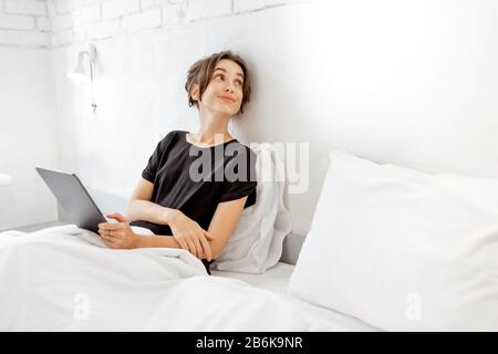 Young woman using digital tablet while sitting relaxed in the bed of a white bedrrom. Concept of a leisure time with mobile wireless devices at home Stock Photo