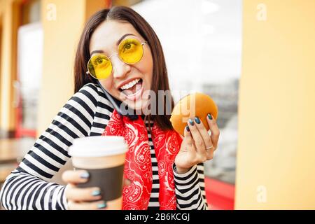 Young woman with smart phone eating burger and drink coffee in fastfood restaurant Stock Photo