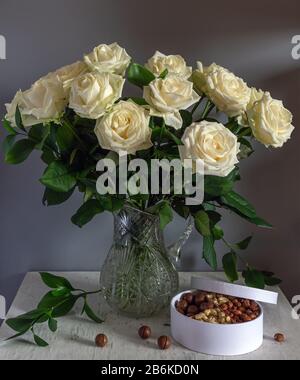 A beautiful bouquet of roses in a crystal jug and a box with tasty nuts. Still life. Vintage. Stock Photo