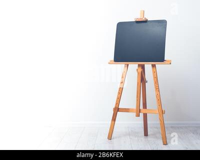 Blank art blackboard, wooden easel, front view, against the background of a white wall. Chalkboard Stock Photo