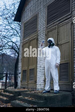Vertical full length snapshot of a scientist standing in front of an old door and observing reaction in glass flask with blue liquid. Ecologist wearing white uniform, gloves and gas mask Stock Photo