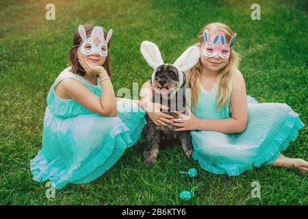 Cute Airedale Terrier dog in Easter bunny ears and little girls in bunny masks on Easter day sitting on grass in garden. Stock Photo
