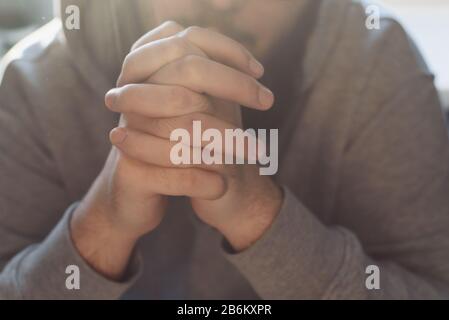 Bearded adult man praying to God sitting at home in the sunbeam. A Muslim or Christian raises his hands to God. Crossed hands in prayer gesture close up. Stock Photo