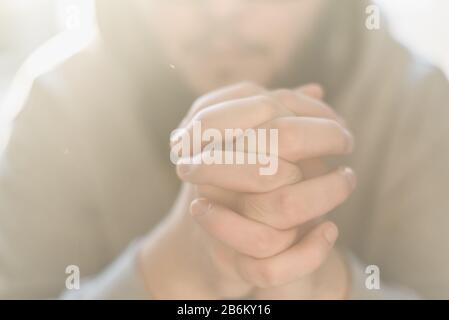 Bearded adult man praying to God sitting at home in the sunbeam. A Muslim or Christian raises his hands to God. Crossed hands in prayer gesture close up. Stock Photo