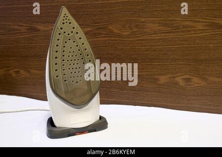 Electric iron with many steam holes on work surface stays vertical on fabric surface on wooden wall background closepu view Stock Photo