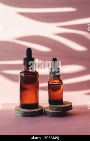 Two glass bottles with moisturizing serum on wooden saw cuts on a pink background. Brown tubes with a dropper on the background of a tropical shadow of a palm leaf. The concept of skin care.