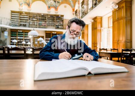 Stylish old senior elderly man with white beard and glasses working in an antique library with books, sitting at the vintage table. Education, library Stock Photo