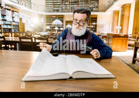 An old intelligent man teacher professor, in stylish casual vintage clothes sitting at the table in ancient library and reading book. Education Stock Photo